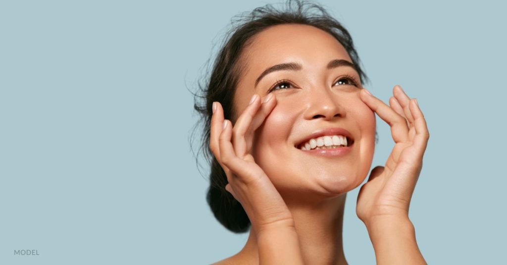 Beautiful woman smiles while looking up and touching face. (Model)