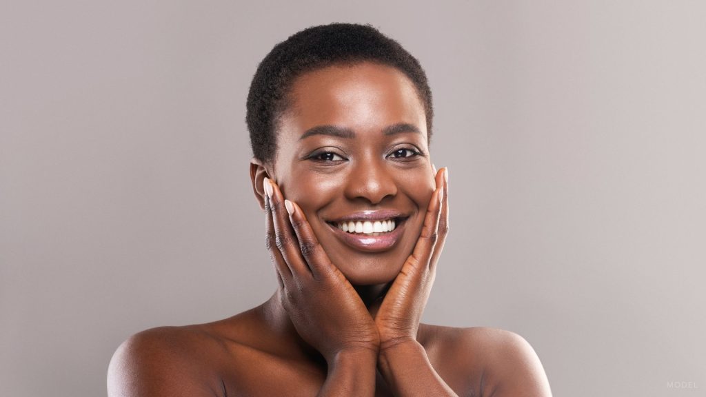A beautiful black woman with glowing skin holds her face while smiling at the camera. (Model)