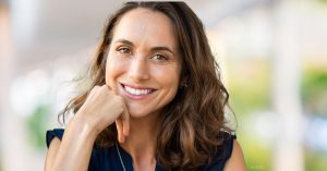 A woman with brown hair is sitting outside with a black shirt on and holding her hand underneath her chin to rest her head. (model)